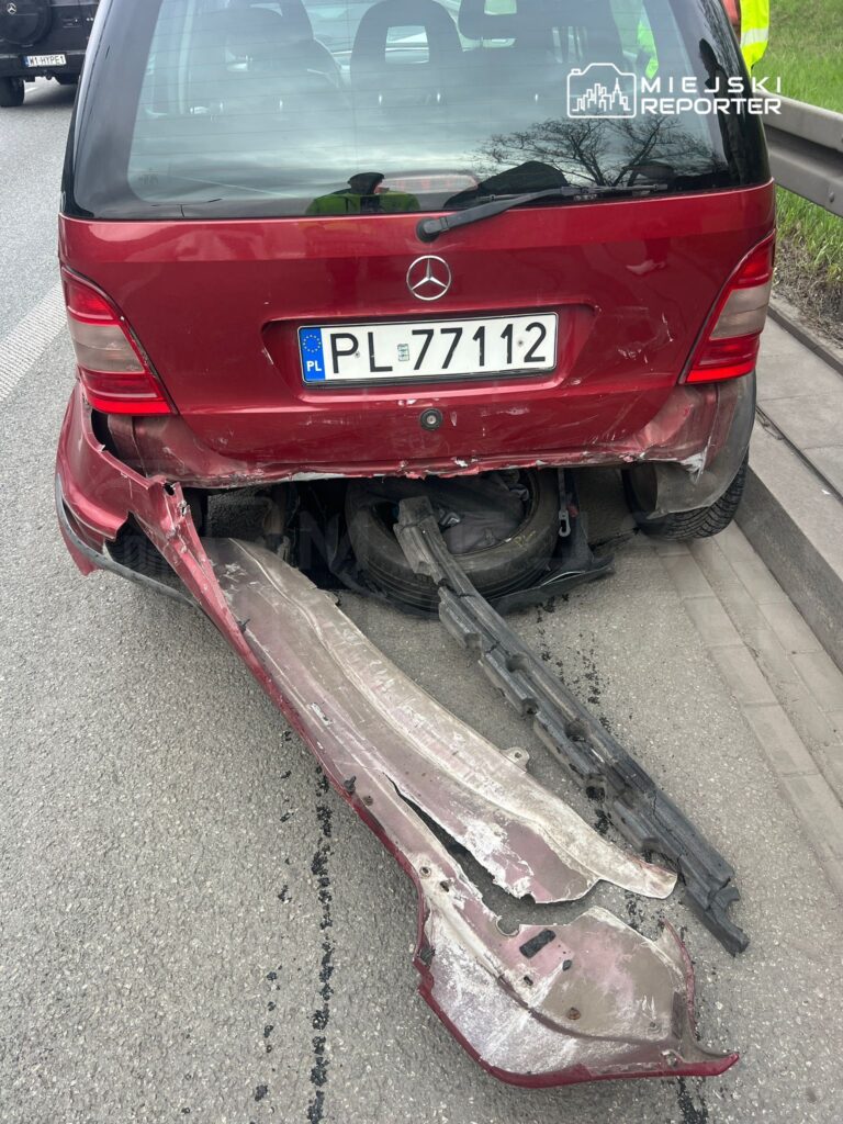 Czerwony samochód osobowy z uszkodzonym tyłem, leżący na poboczu drogi, obok odłamki zderzaka.