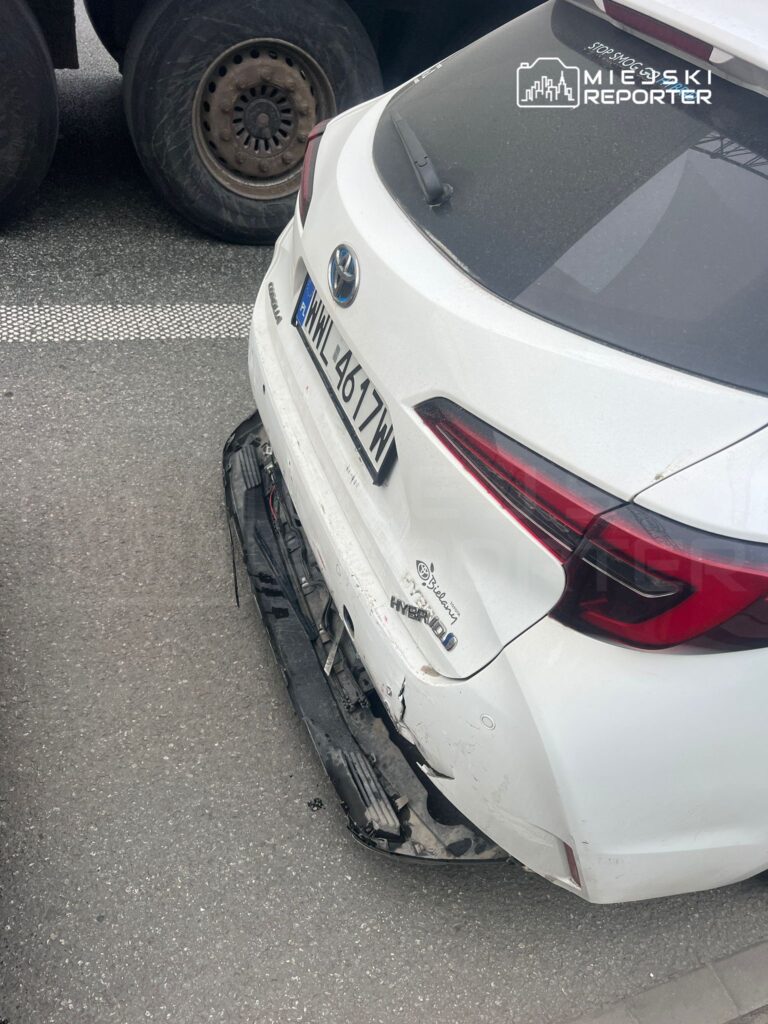 Uszkodzona biała Toyota z widocznymi zarysowaniami na tylnej części zaparkowana obok dużego pojazdu ciężarowego.