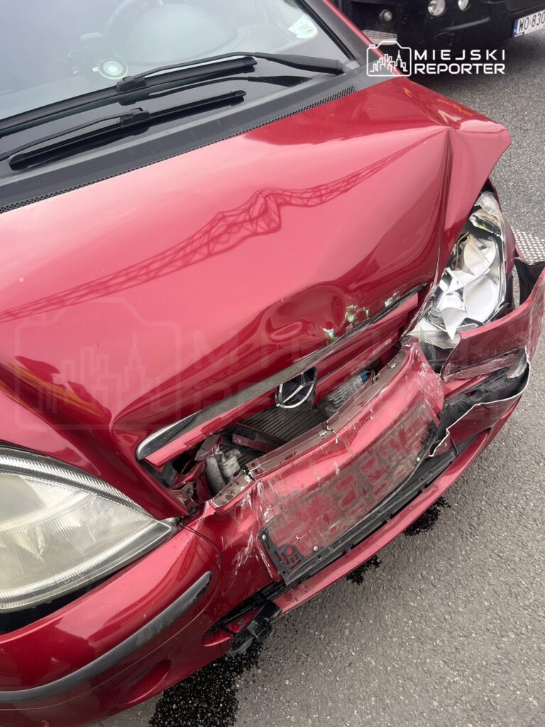 Czerwony samochód osobowy z uszkodzoną maską i zderzakiem, zaparkowany na drodze.