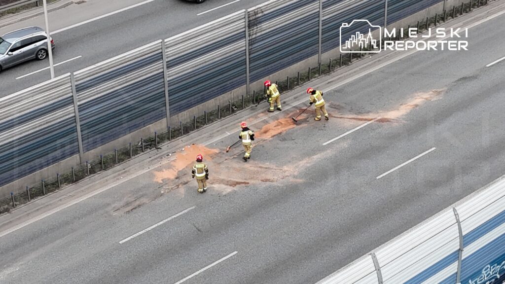 Czterech strażaków w żółtych mundurach usuwa plamę na jezdni, obok ruchliwej drogi.