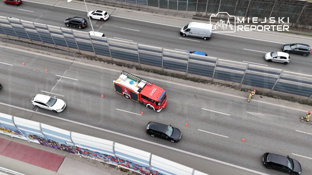 Czerwony wóz strażacki zatrzymany na jezdni, obok dwóch strażaków w mundurach, ustawiających pachołki.