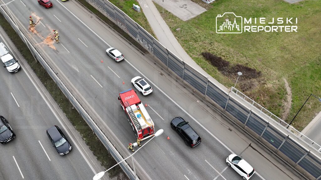 Strażacy oczyszczają jezdnię z plamy substancji na ruchliwej drodze, obok przejeżdżające samochody.