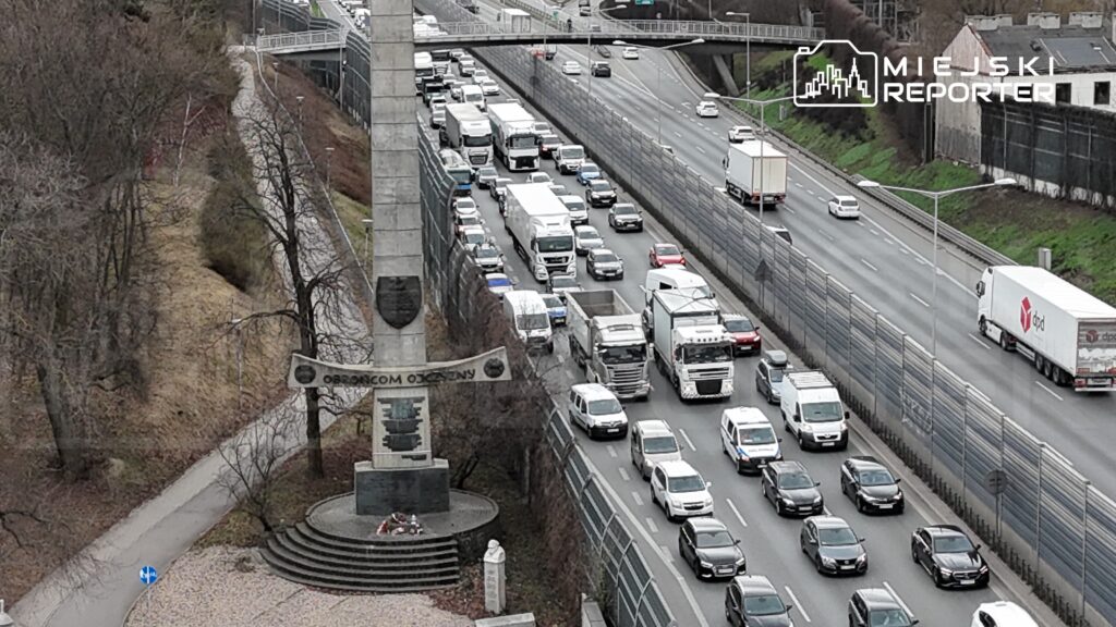 Zator drogowy z ciężarówkami i samochodami osobowymi na ruchliwej jezdni obok pomnika z napisem "OBROŃCOM OJCZYZNY".
