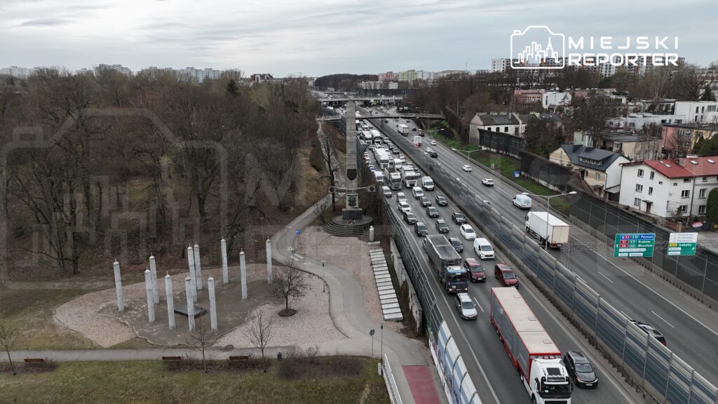 Ruchliwa ulica z samochodami, obok pomnik z kolumnami i alejkami w parku, w tle bloki mieszkalne.