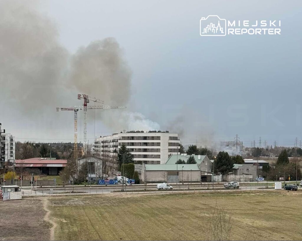 W tle widoczny dym unoszący się z budowy obok nowoczesnego osiedla mieszkaniowego, w otoczeniu terenów zielonych.
