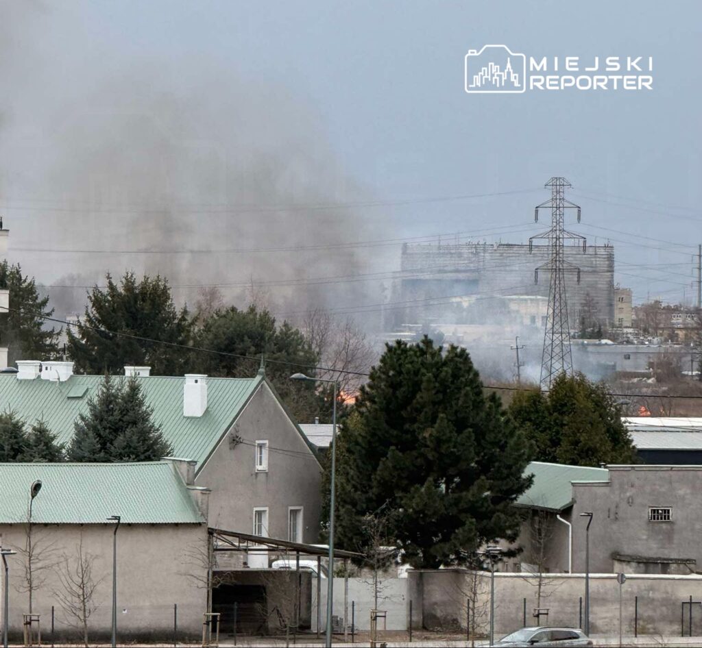 Gęsty dym unoszący się nad budynkami mieszkalnymi w miejskim krajobrazie, w tle widoczna konstrukcja budowlana.