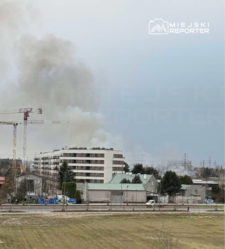 Gęsty dym unoszący się nad nowoczesnym budynkiem mieszkalnym w otoczeniu placu budowy i zieleni.