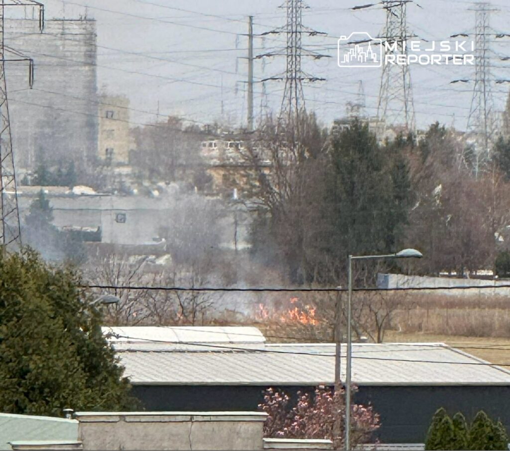 W oddali widać dym wydobywający się z płonącej trawy na łące, otoczonej budynkami i słupami energetycznymi.