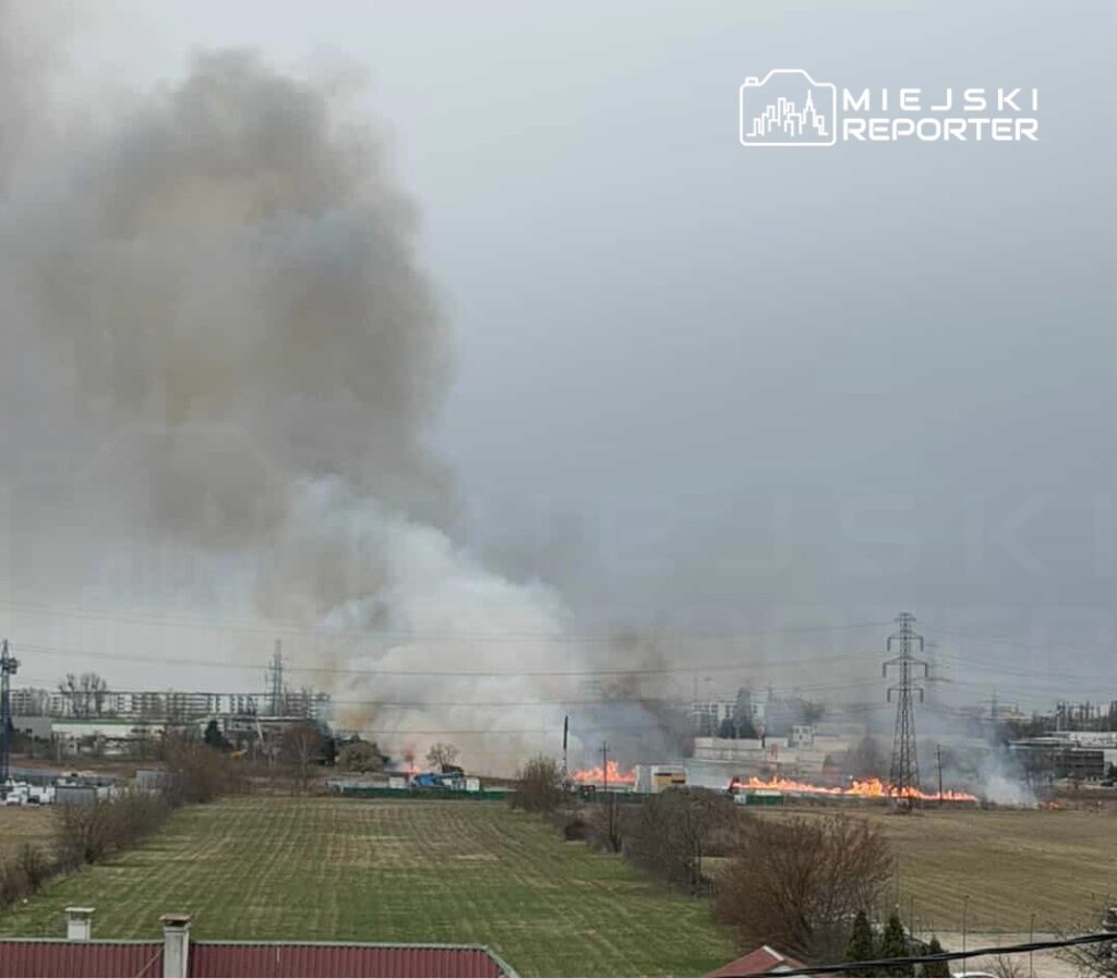 Gęsty dym unoszący się nad płonącym terenem w pobliżu zabudowań przemysłowych, z widocznymi płomieniami.