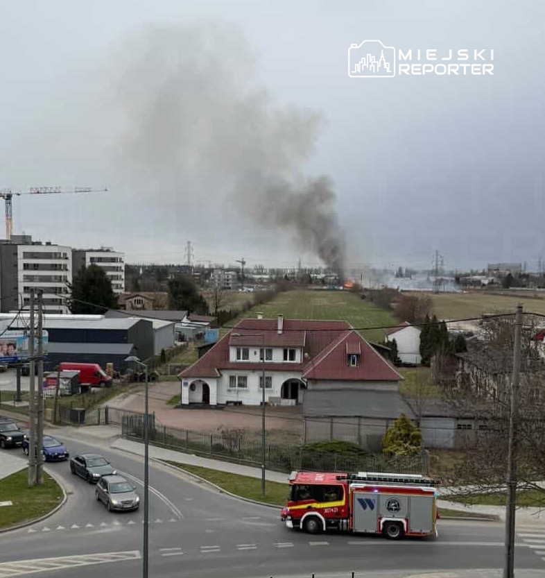 Czarny dym unosi się nad polem, w tle widać płonący obiekt, a w kierunku zdarzenia jedzie wóz strażacki.