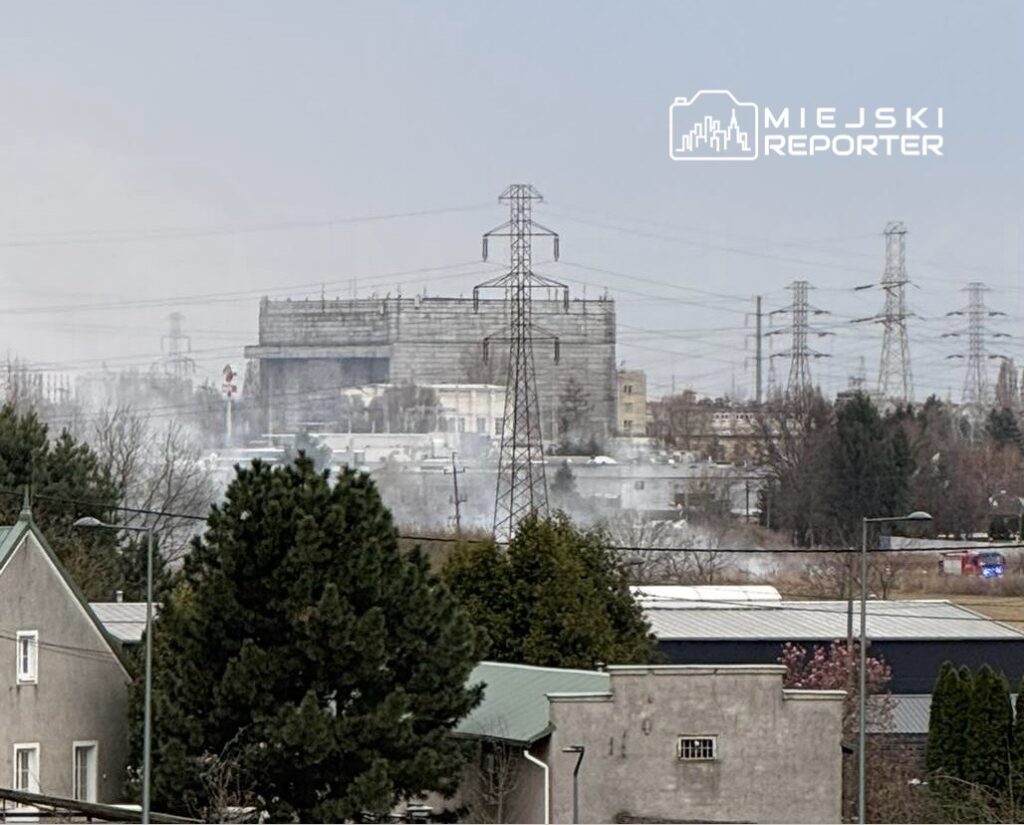 Widok na przemysłowy obszar z dymem unoszącym się w powietrzu, w tle widać wysokie słupy energetyczne.