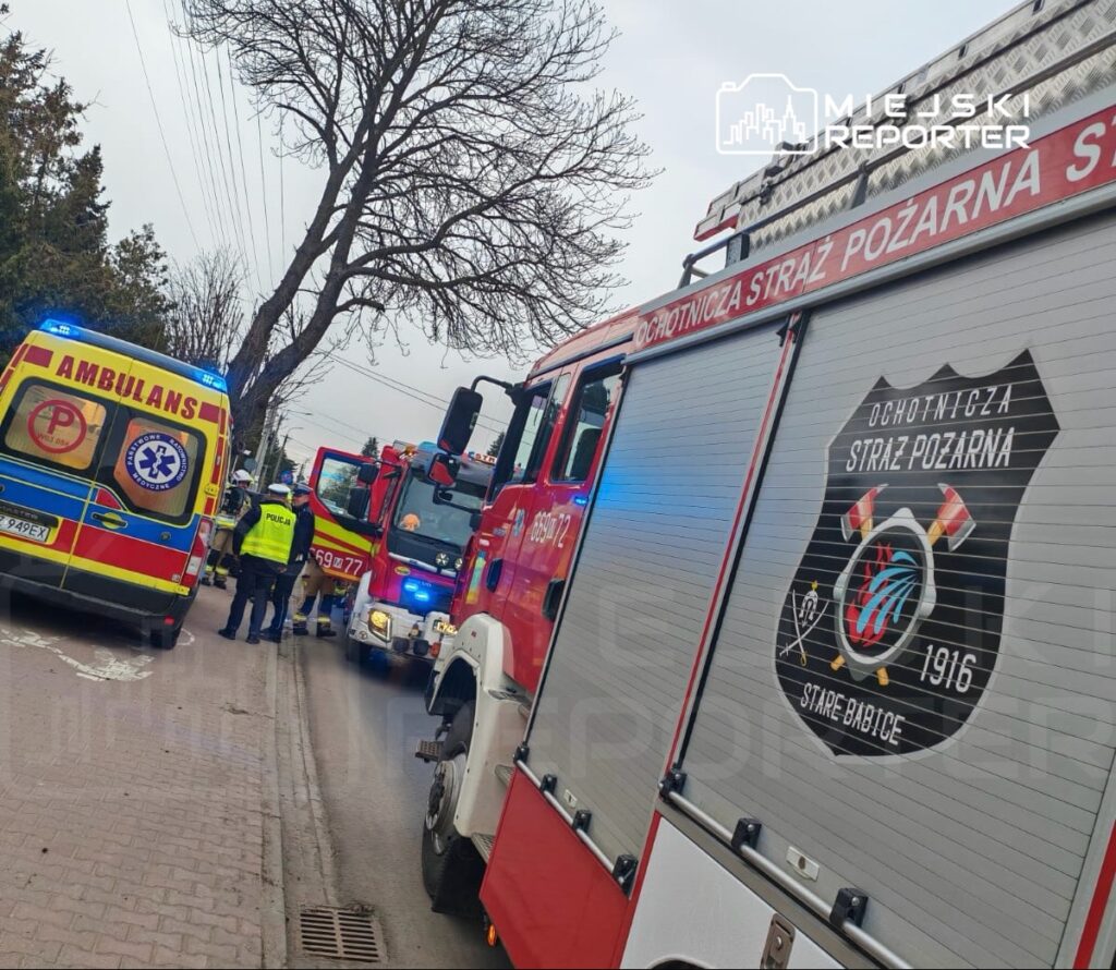 Zespół ratunkowy w akcji przy wozach strażackich i ambulansie na ulicy, w tle drzewa i budynki.