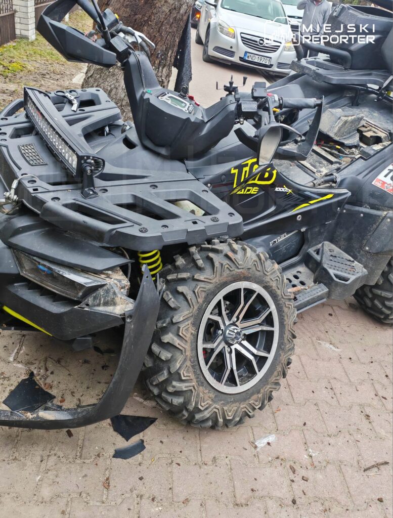 Quad z uszkodzoną przednią częścią i dużą oponą stoi na chodniku, obok zaparkowanych samochodów.