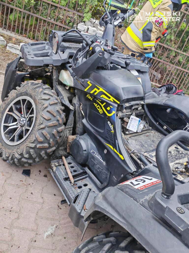 Uszkodzony quad leży na chodniku, obok niego widać fragmenty zniszczonego pojazdu oraz osobę w mundurze strażaka.