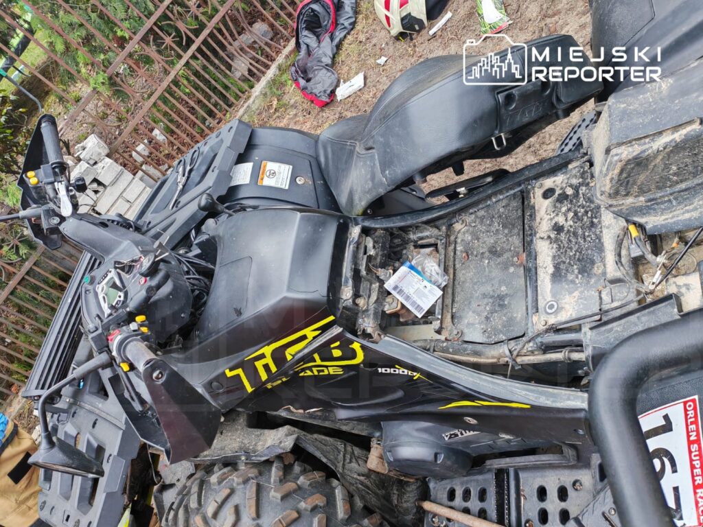 Czarny quad z widocznymi elementami sterującymi, leży na ziemi w nieutwardzonym otoczeniu, obok porzucone przedmioty.