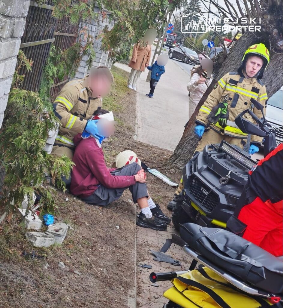 Strażak udziela pomocy poszkodowanemu mężczyźnie siedzącemu na chodniku, obok stoi ambulans. W tle przechodnie i ruch uliczny.