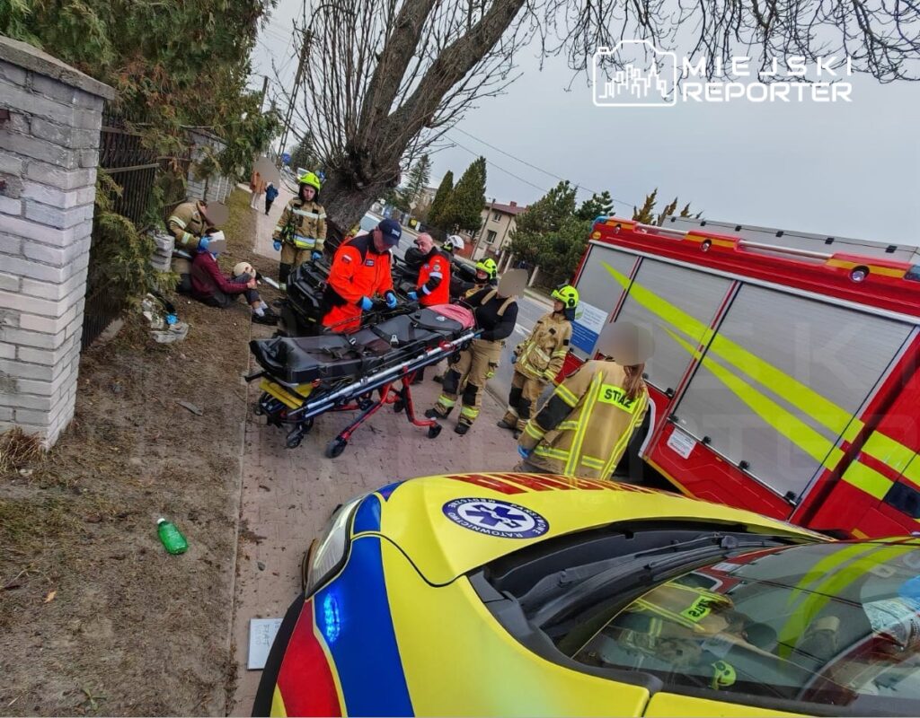 Grupa ratowników medycznych i strażaków udziela pomocy osobie siedzącej na chodniku obok karetki i wozu strażackiego.