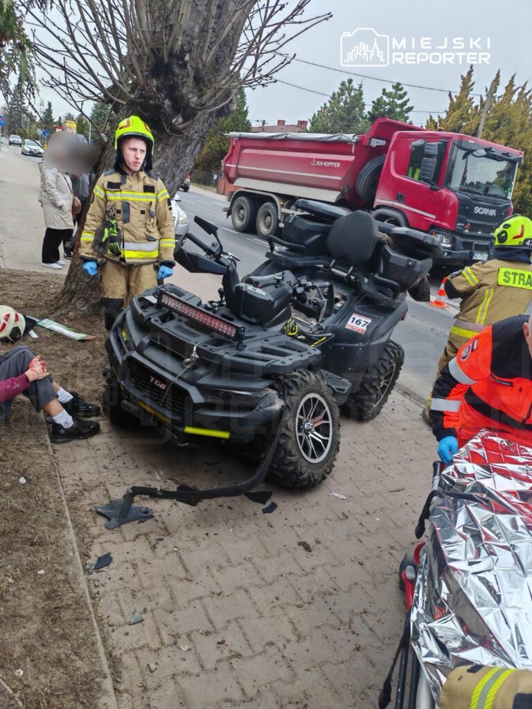 Strażak w hełmie obserwujący uszkodzony quad na chodniku, obok poszkodowany mężczyzna z raną na nodze.