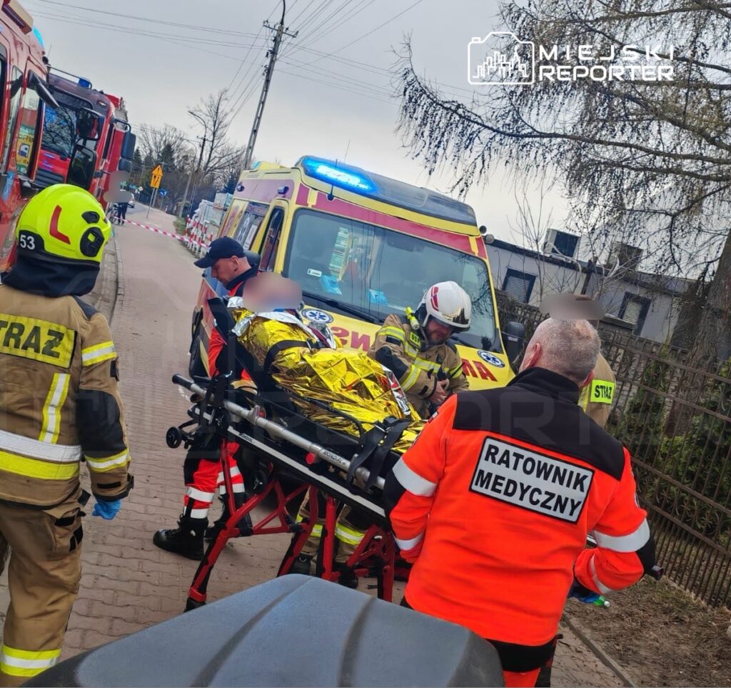 Ratownicy medyczni i strażacy udzielają pomocy poszkodowanemu na noszach, obok karetki na ulicy.