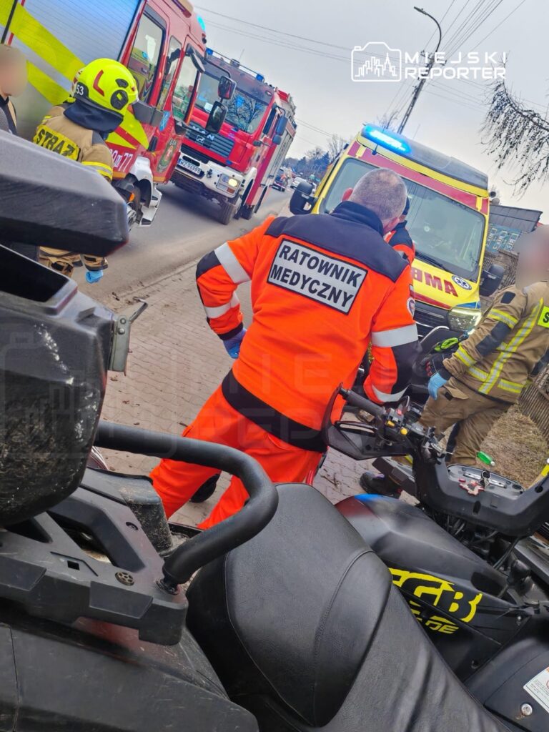 Ratownik medyczny w pomarańczowym uniformie rozmawia z innymi służbami na miejscu zdarzenia, obok wozu strażackiego i karetki.