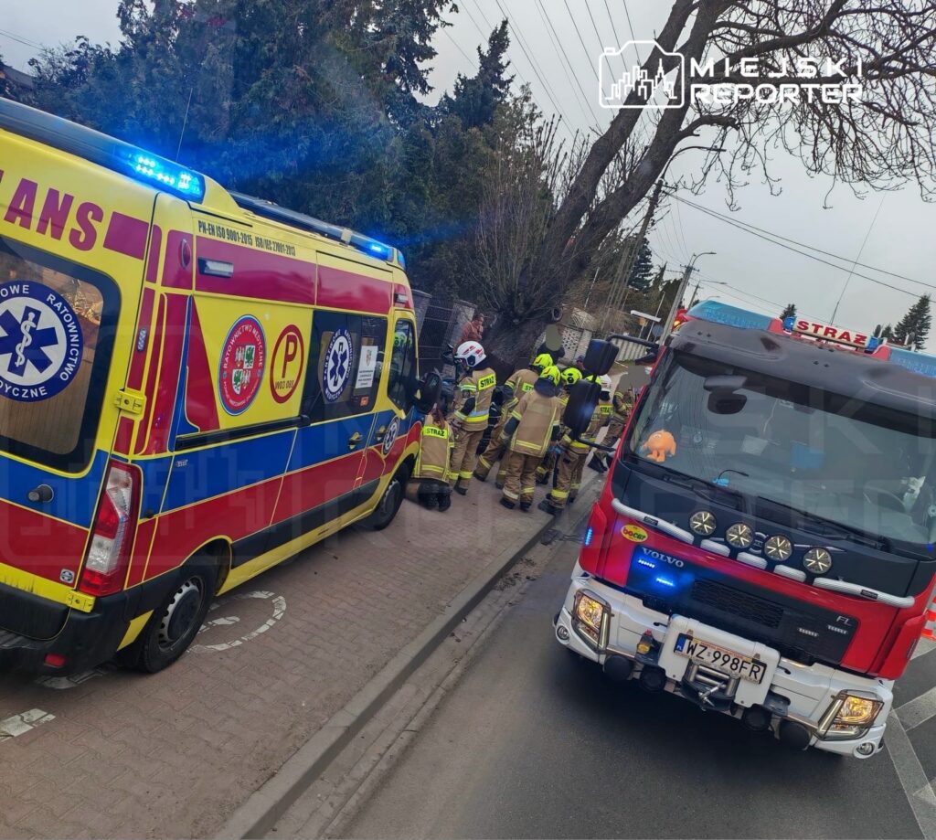 Zespół ratunkowy i strażacy udzielają pomocy przy wypadku drogowym, obok dwóch pojazdów ratunkowych.