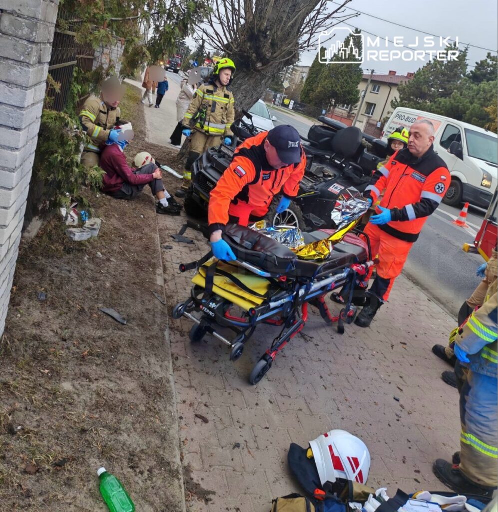 Ratownicy medyczni przygotowują nosze dla poszkodowanego na chodniku, w otoczeniu strażaków i pojazdów ratunkowych.