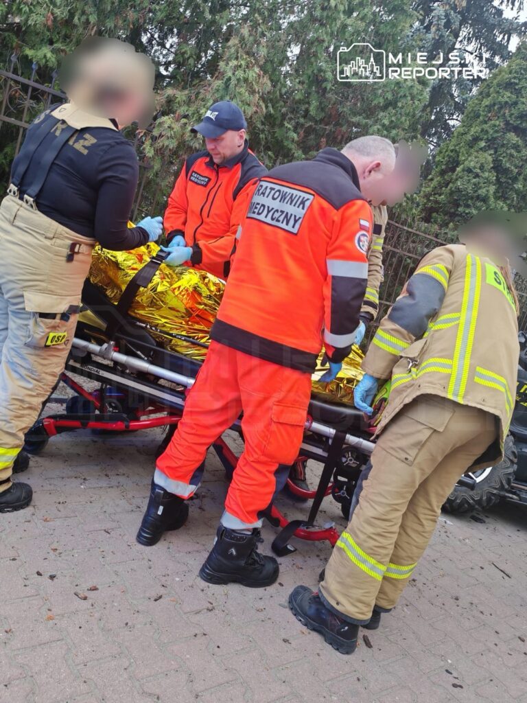 Grupa ratowników medycznych i strażaków udziela pomocy poszkodowanemu leżącemu na noszach w terenie zadrzewionym.