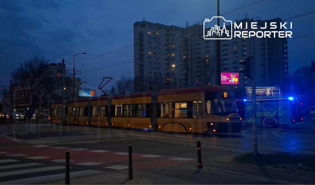 Tramwaj zatrzymany na skrzyżowaniu, w tle widoczny budynek mieszkalny oraz radiowóz z włączonymi sygnałami świetlnymi.
