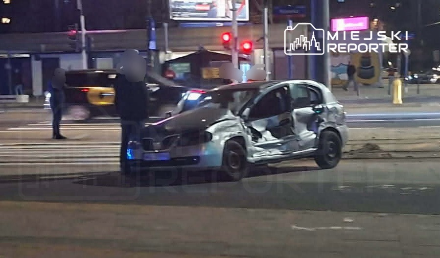 Uszkodzony samochód osobowy stoi na skrzyżowaniu, obok grupy ludzi rozmawiających z policjantem.