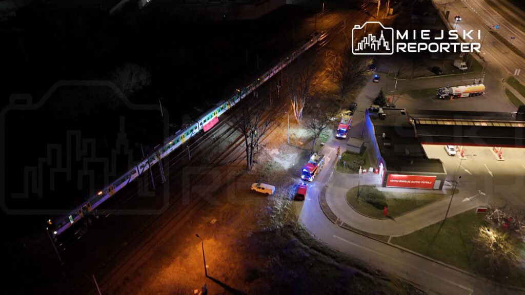 Pociąg pasażerski przejeżdża obok stacji, przy której znajdują się pojazdy straży pożarnej oraz zaparkowana ciężarówka.