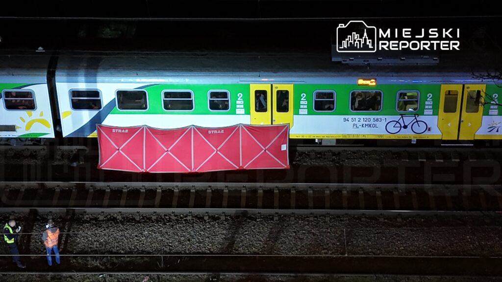 Na torach stoi pociąg z kolorową grafiką, a obok dwóch mężczyzn w kamizelkach odblaskowych rozmawia przy czerwonym parawanie.