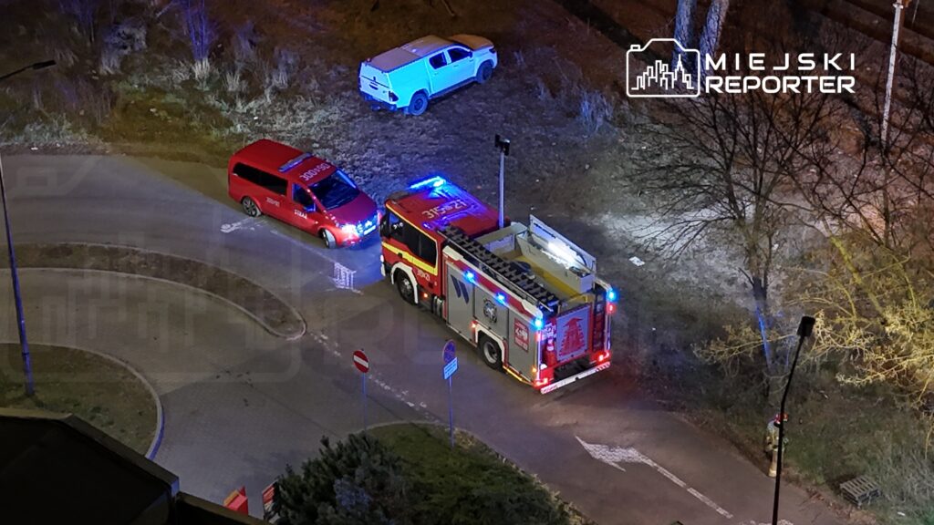 W nocy na osiedlowej drodze zaparkowane są pojazdy ratunkowe: wóz strażacki z włączonymi sygnałami świetlnymi oraz oznakowany samochód ratunkowy.