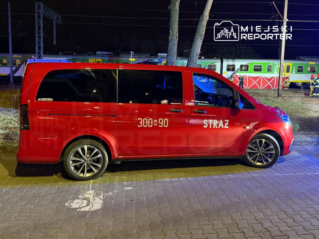 Czerwony samochód strażacki zaparkowany na kostce brukowej obok torów kolejowych w nocy.