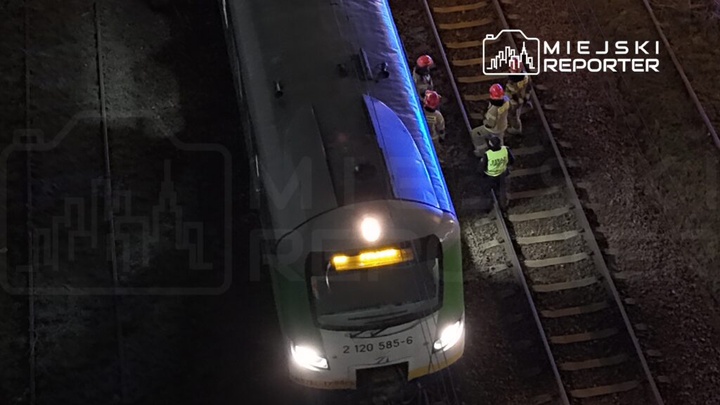 Grupa pracowników w kaskach omawia sytuację obok pociągu na torach kolejowych w nocy.