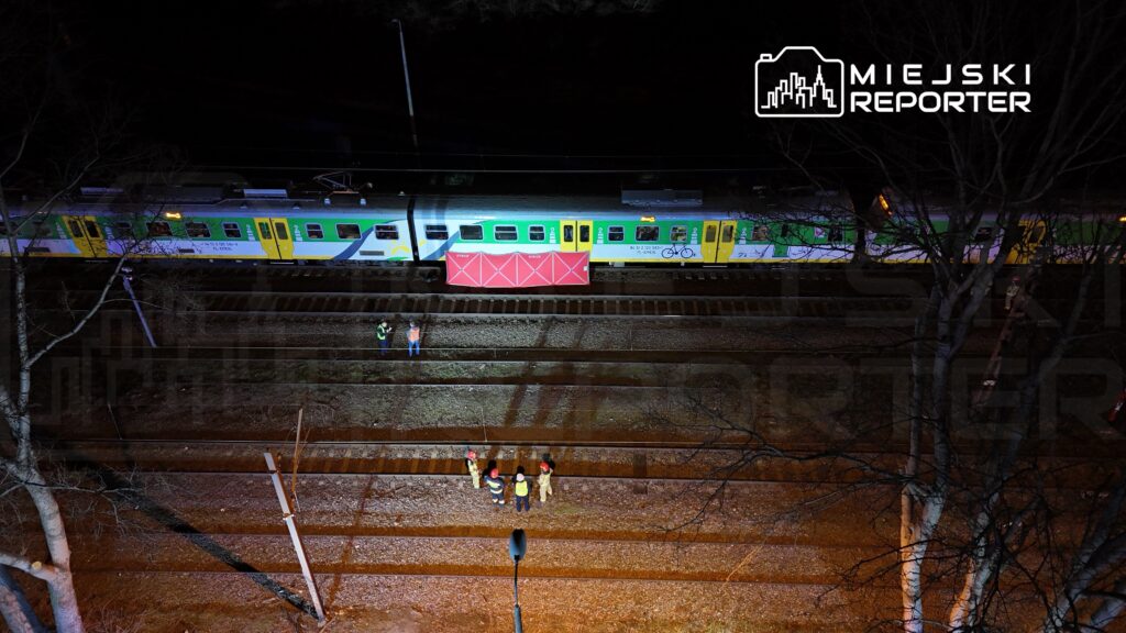 Grupa pracowników w odblaskowych kamizelkach prowadzi działania obok pociągu na torach w nocy.