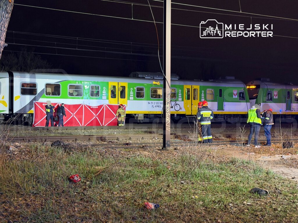 Grupa ratowników w kamizelkach odblaskowych zabezpiecza miejsce wypadku kolejowego obok pociągu na torach.