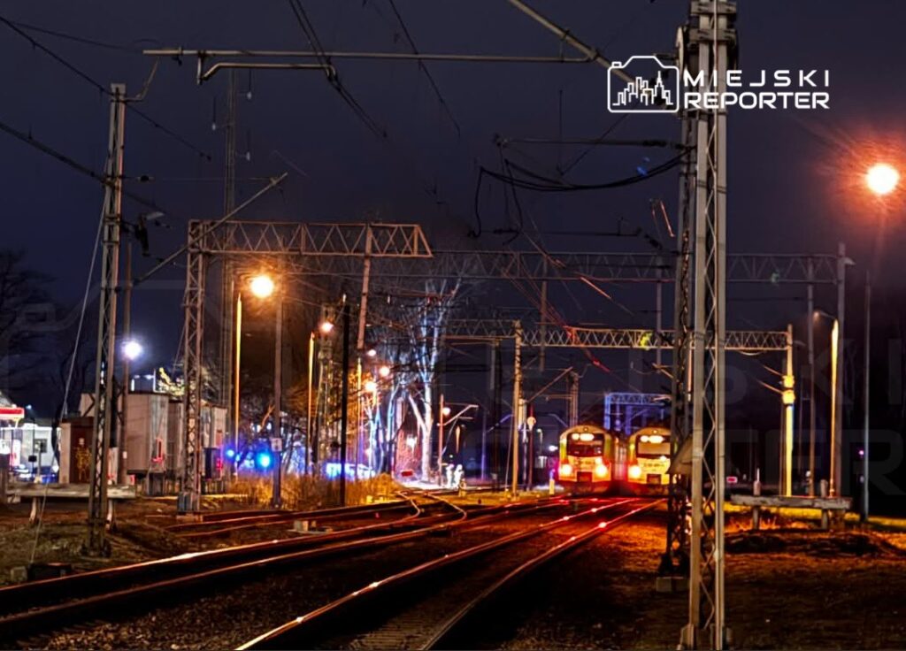 Dwa pociągi stoją na torach w oświetlonym nocy otoczeniu stacji kolejowej, z widocznymi latarniami i siecią trakcyjną.