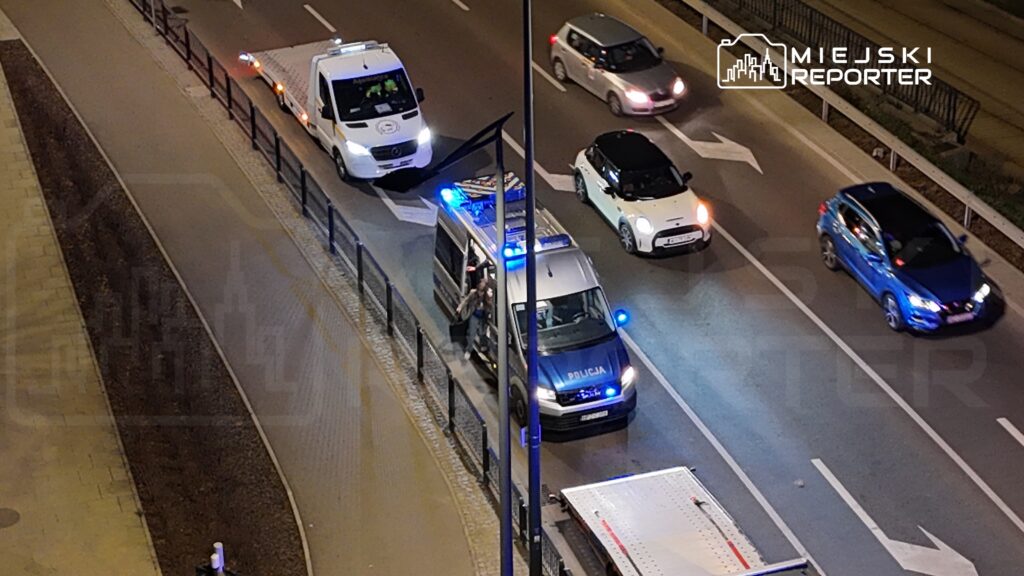 Policyjny radiowóz z włączonymi sygnałami świetlnymi zatrzymuje się na ulicy, obok niego stoi mężczyzna z torbą.