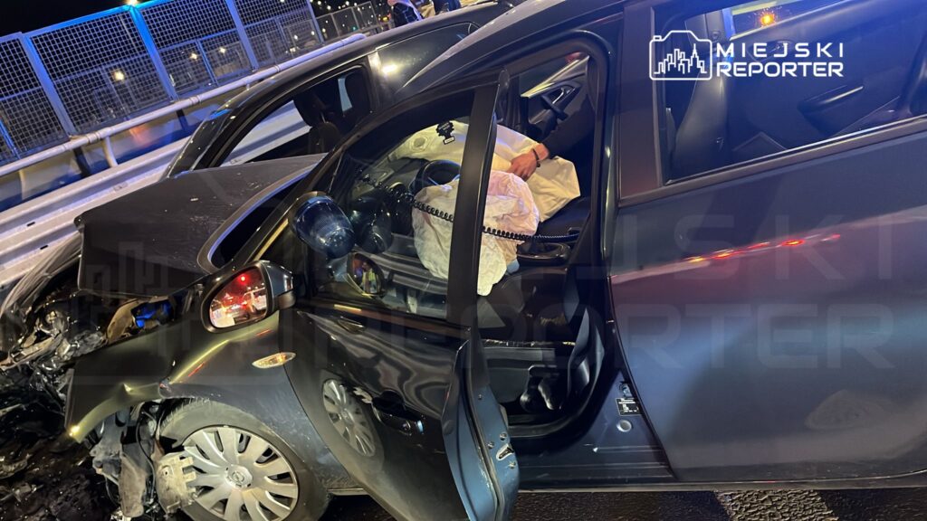 Zniszczony samochód osobowy z otwartymi drzwiami, w którym widoczna jest poduszka powietrzna, na drodze nocą.
