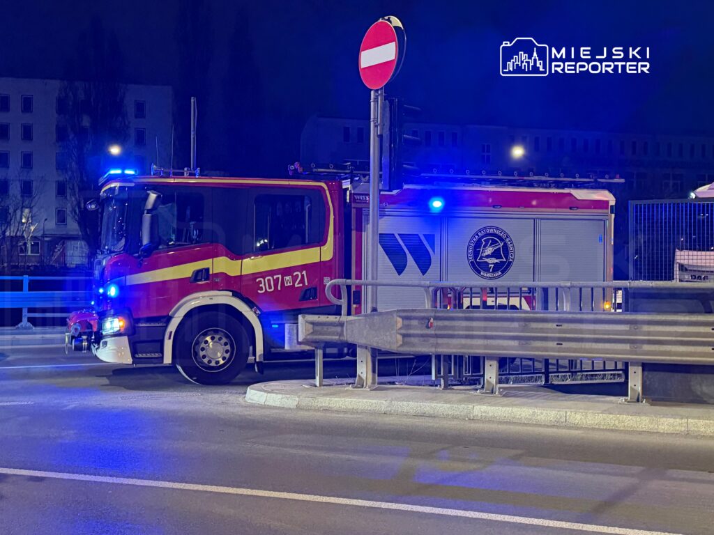 Wozidło strażackie z niebieskimi światłami zatrzymane na ulicy w nocy, obok znaku drogowym zakazu wjazdu.