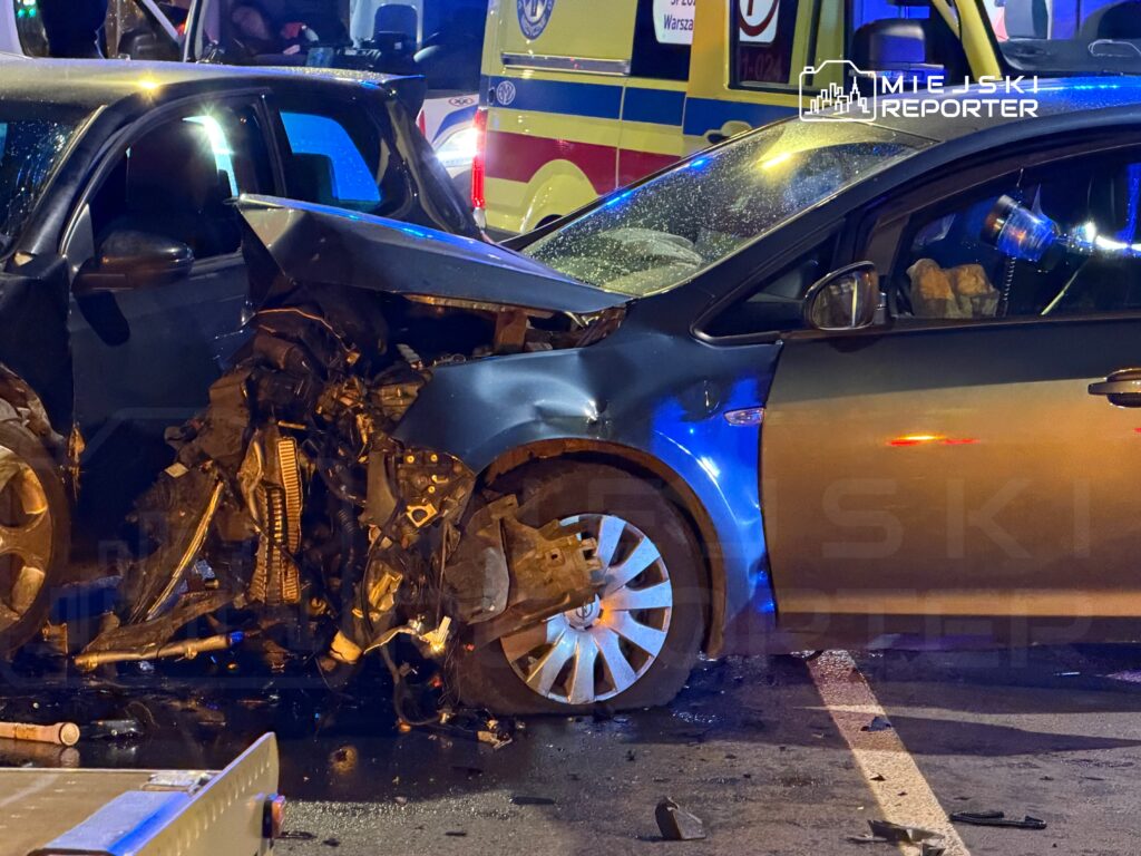 Dwa uszkodzone samochody po zderzeniu na ulicy, w tle widać radiowóz i karetkę.