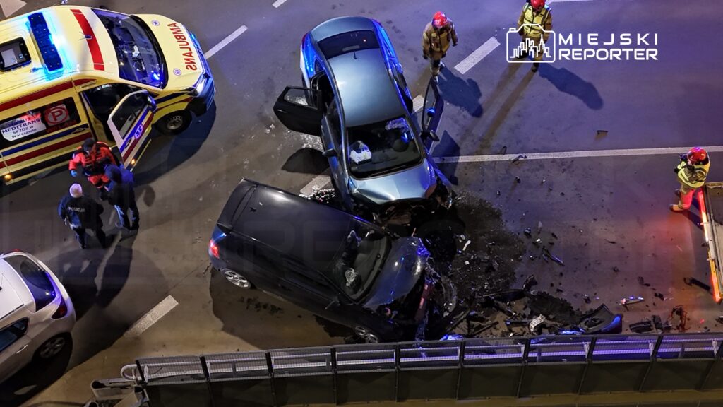 Dwa pojazdy osobowe zderzone na jezdni, otoczone przez zespół ratunkowy i strażaków, w nocy.