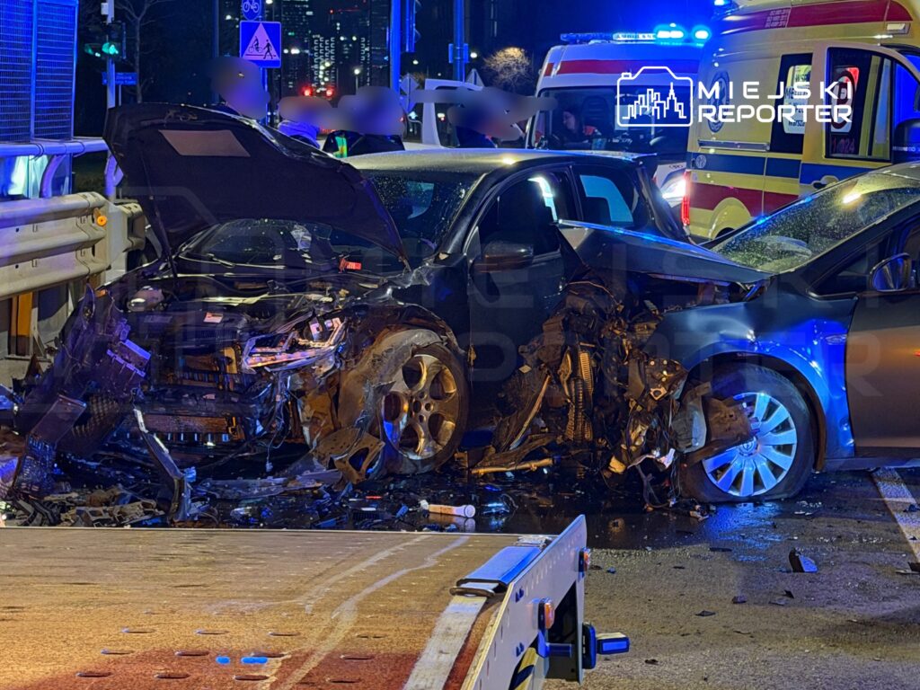 Zniszczony samochód osobowy z uszkodzoną przednią częścią stoi na drodze, obok widać pojazdy służb ratunkowych.