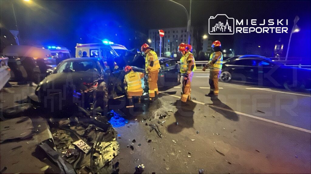 Grupa ratowników w odblaskowych uniformach bada wrak samochodu po wypadku drogowym na oświetlonej ulicy.