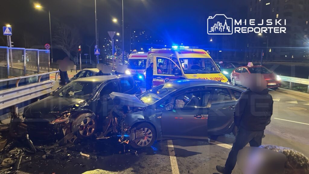 Dwa uszkodzone pojazdy po kolizji na miejskiej ulicy, obok stojące karetki i kilka osób obserwujących zdarzenie.