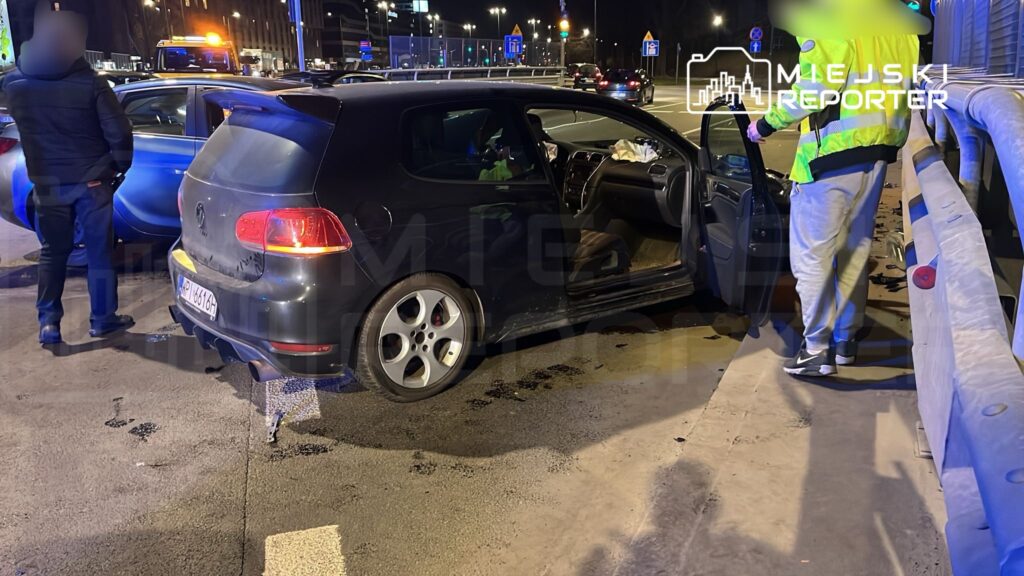 Czarny samochód osobowy stoi na parkingu, obok dwóch mężczyzn, jeden w odblaskowej kamizelce, rozmawiających.