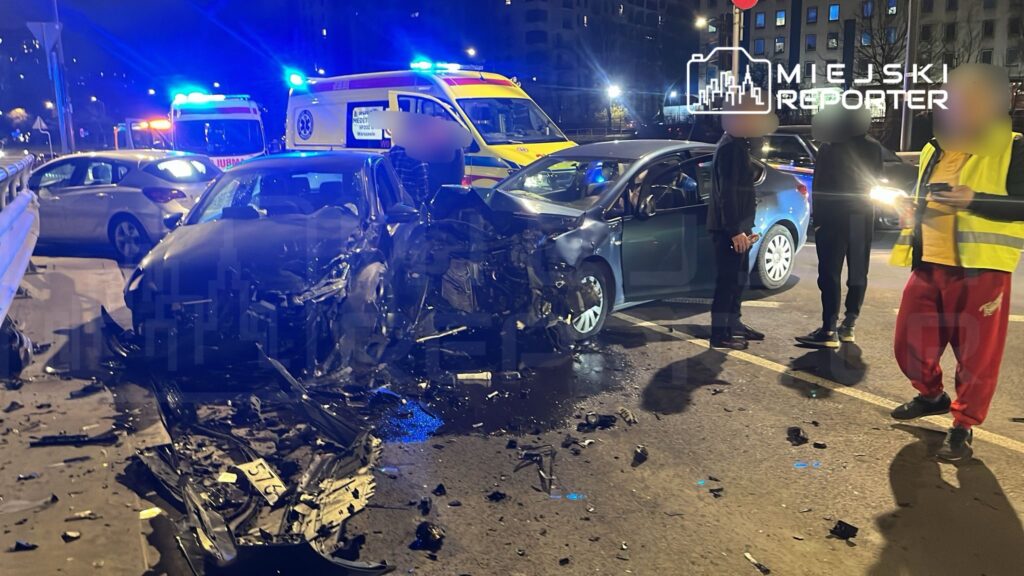 Dwa uszkodzone samochody po wypadku drogowym, obok stoją osoby oraz pojazdy służb ratunkowych z włączonymi sygnałami.