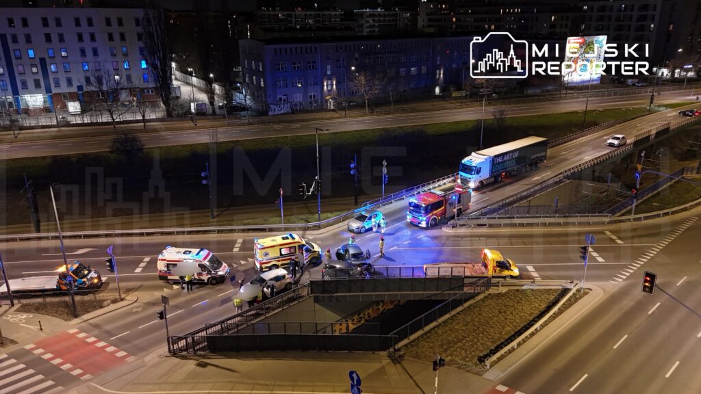 W nocy na skrzyżowaniu ulic stoją pojazdy służb ratunkowych oraz policji, a w tle widać oświetlone budynki.