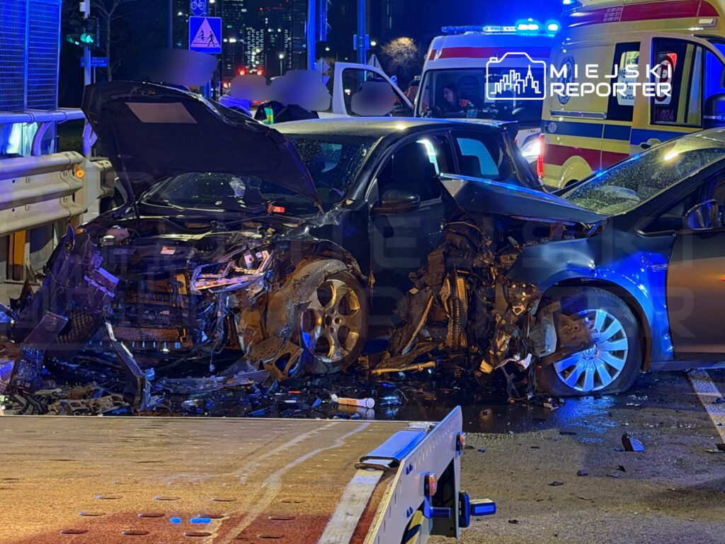 Zniszczony samochód osobowy z otwartą maską stoi na ulicy, obok widać pojazdy służb ratunkowych z włączonymi sygnałami.
