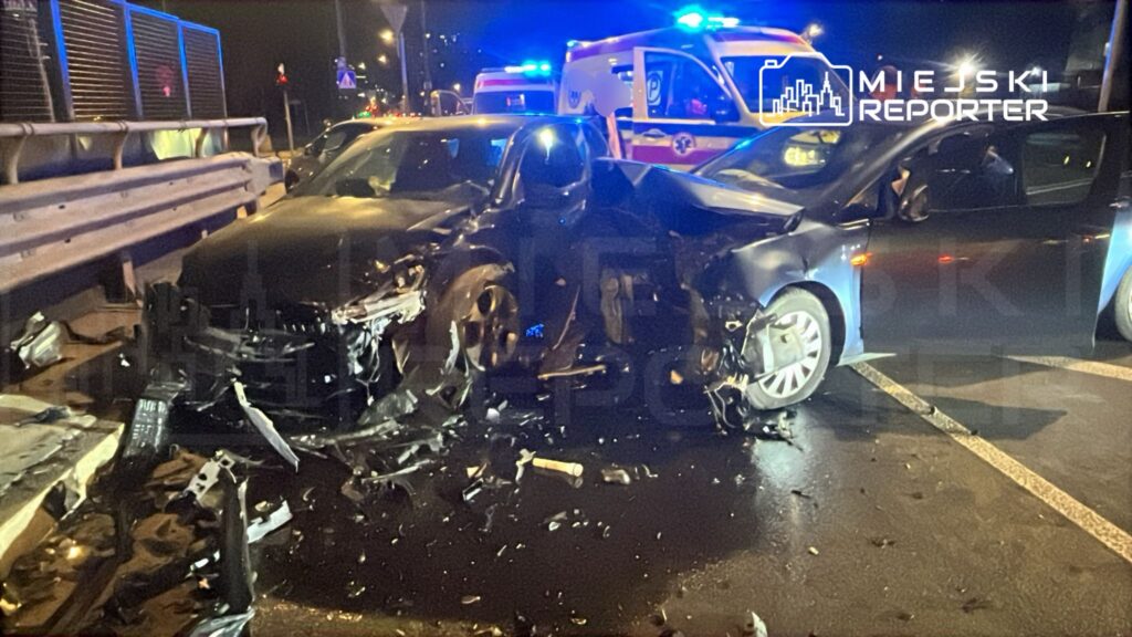 Dwa uszkodzone samochody po wypadku drogowym na ulicy, w tle widać radiowóz z włączonymi sygnałami świetlnymi.